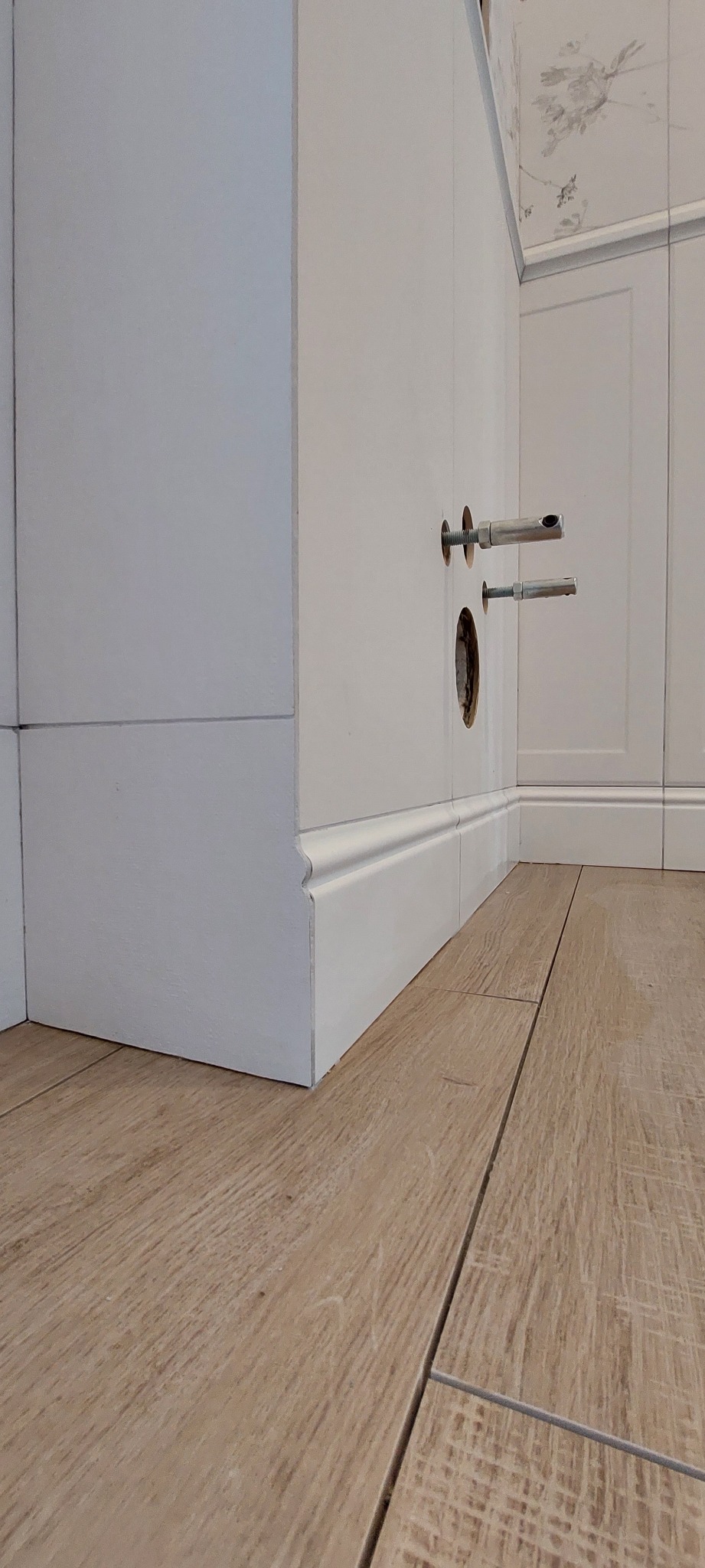 Fragment ściany z dwiema dziurami i kołkami rozporowymi, biała listwa przypodłogowa i podłoga z paneli imitujących drewno, widoczne płytki na ścianie w tle.