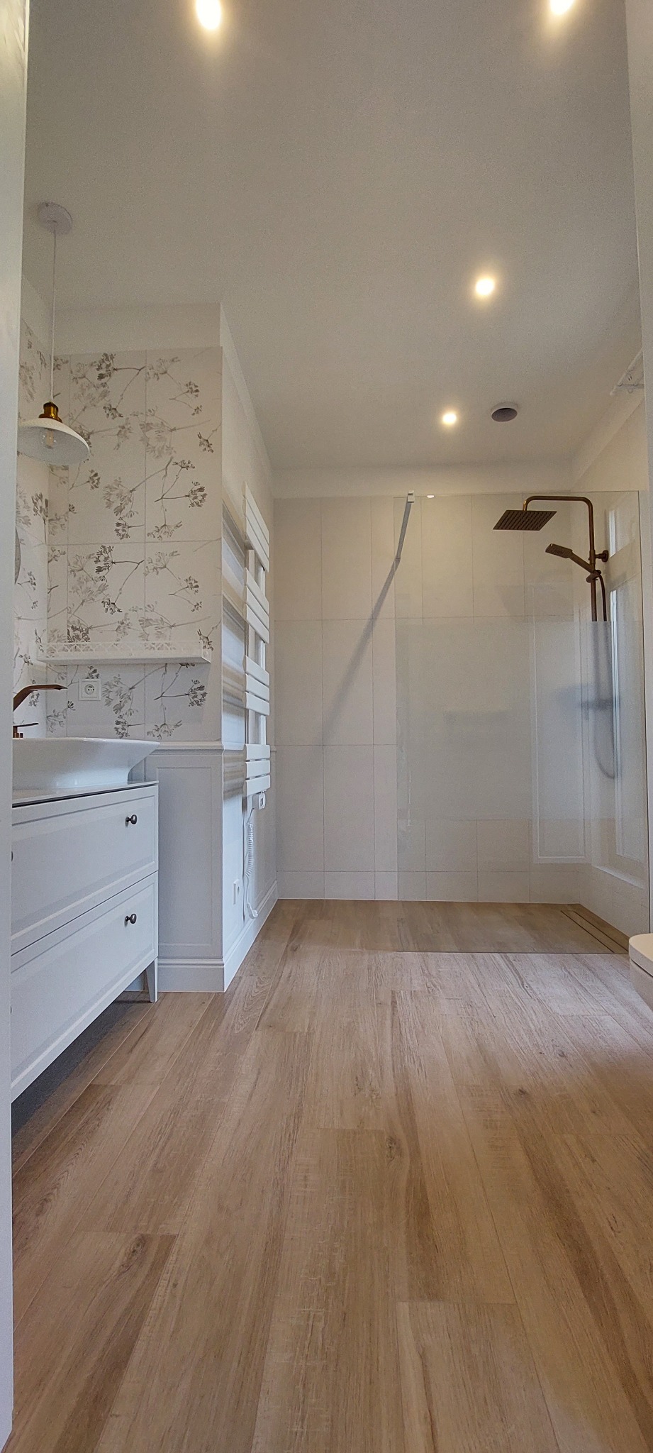 Jasna łazienka z drewnopodobną podłogą, białą armaturą, kabiną prysznicową typu walk-in i grzejnikiem drabinkowym, wykończona płytkami w kwiatowy wzór.