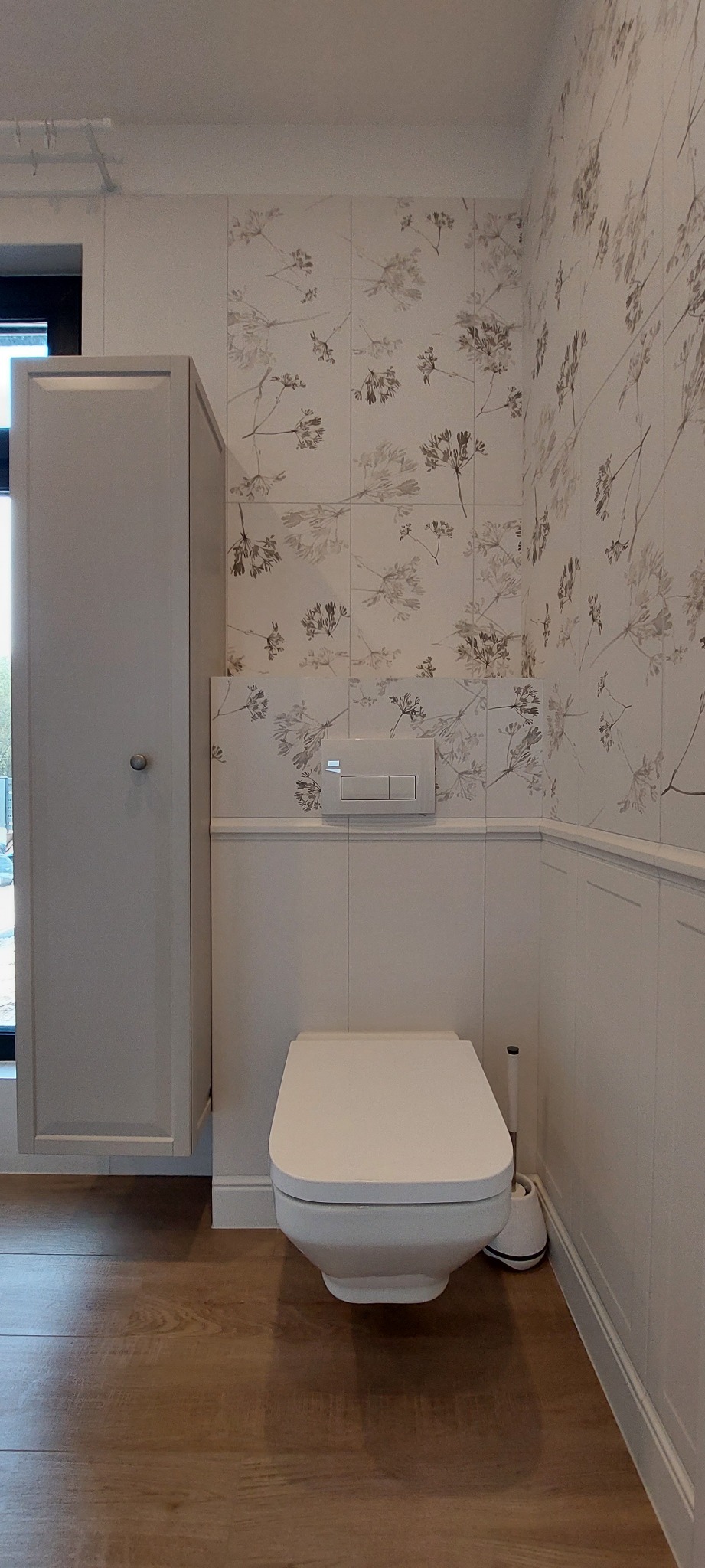 Nowoczesna toaleta wisząca w łazience z drewnopodobną podłogą, białą szafką i płytkami w kwiatowe wzory.