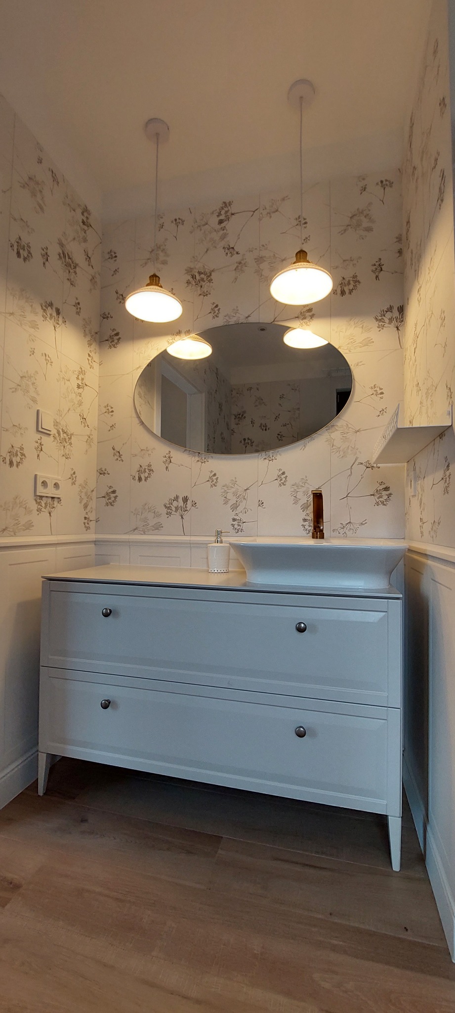 Nowoczesna łazienka z umywalką nablatową na szarej szafce z dwoma szufladami, owalne lustro i trzy okrągłe lampy wiszące, ściany wyłożone płytkami w kwiatowy wzór.