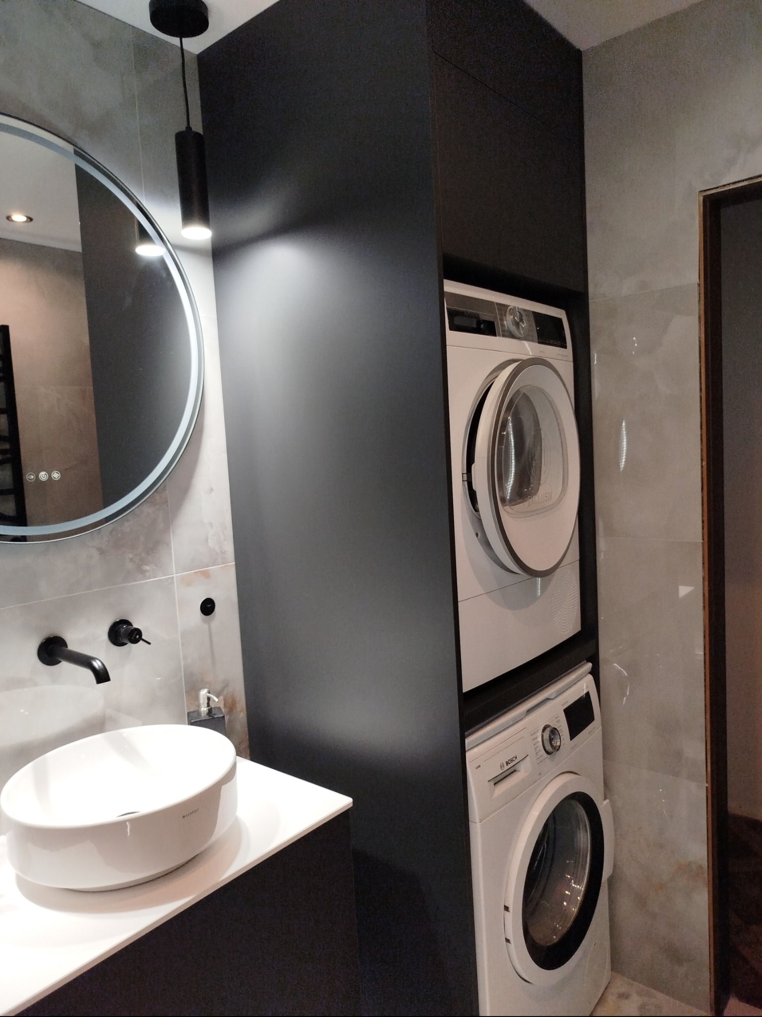 Nowoczesna łazienka z umywalką nablatową, okrągłym lustrem z podświetleniem LED i zabudową z pralką i suszarką w kolorze czarnym, wykończona płytkami imitującymi marmur.