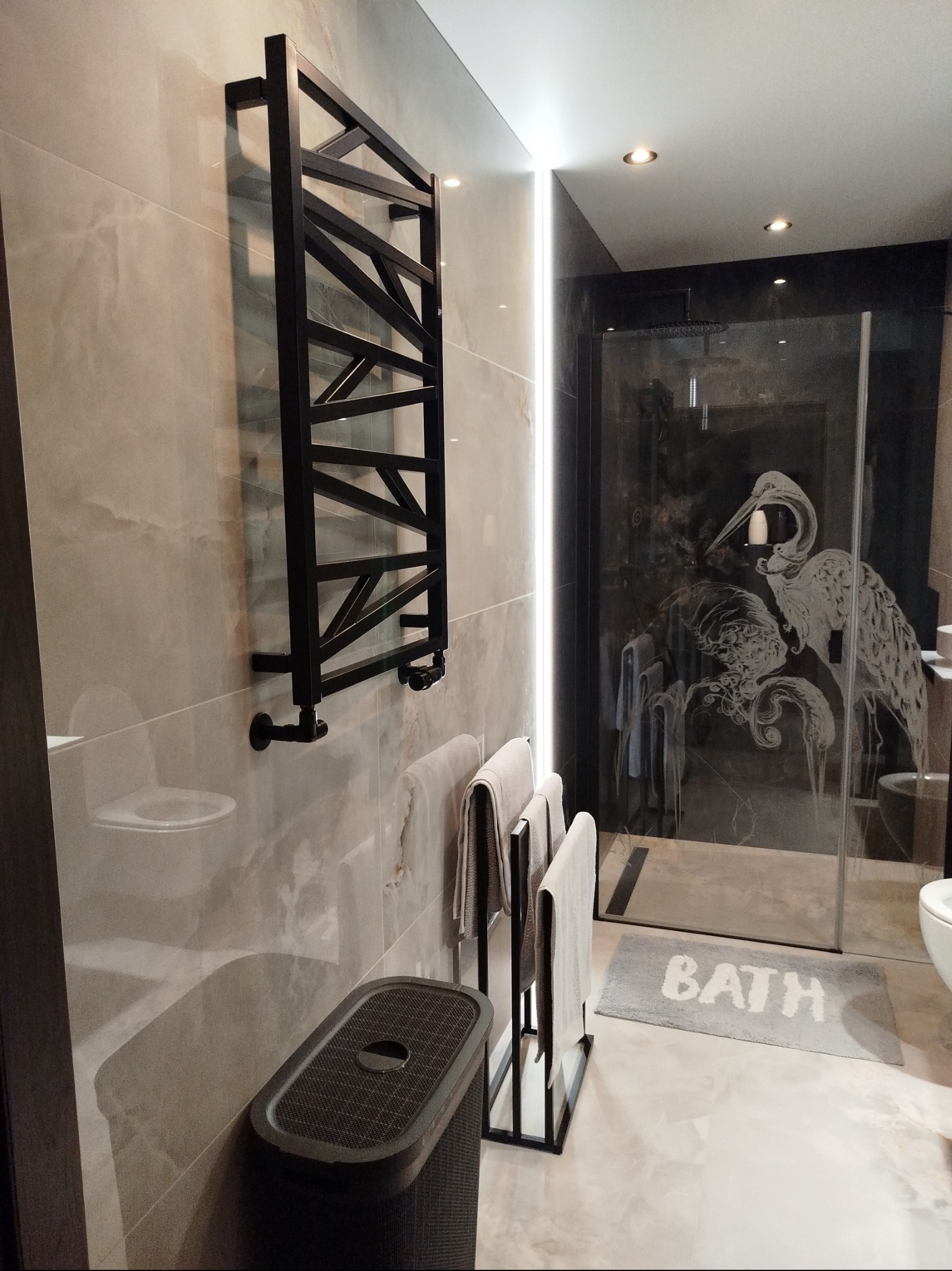 Nowoczesna łazienka z czarnym grzejnikiem drabinkowym, szklaną kabiną prysznicową z motywem ptaków i beżowymi płytkami. Minimalistyczny design i subtelne oświetlenie.