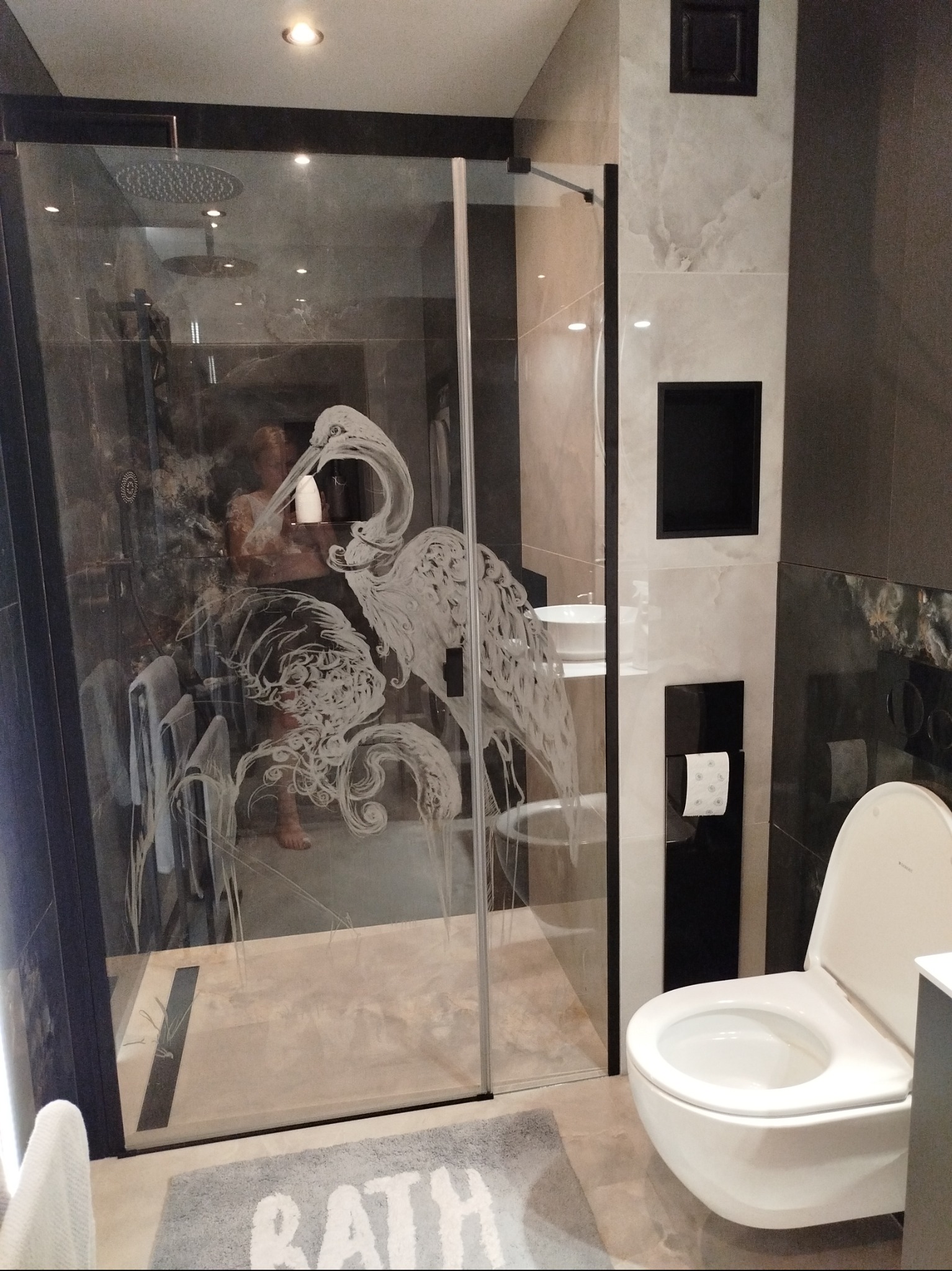 Nowoczesna łazienka z kabiną prysznicową zdobioną motywem żurawi, widoczna toaleta i elementy wykończenia w odcieniach szarości i bieli. Minimalistyczny design.