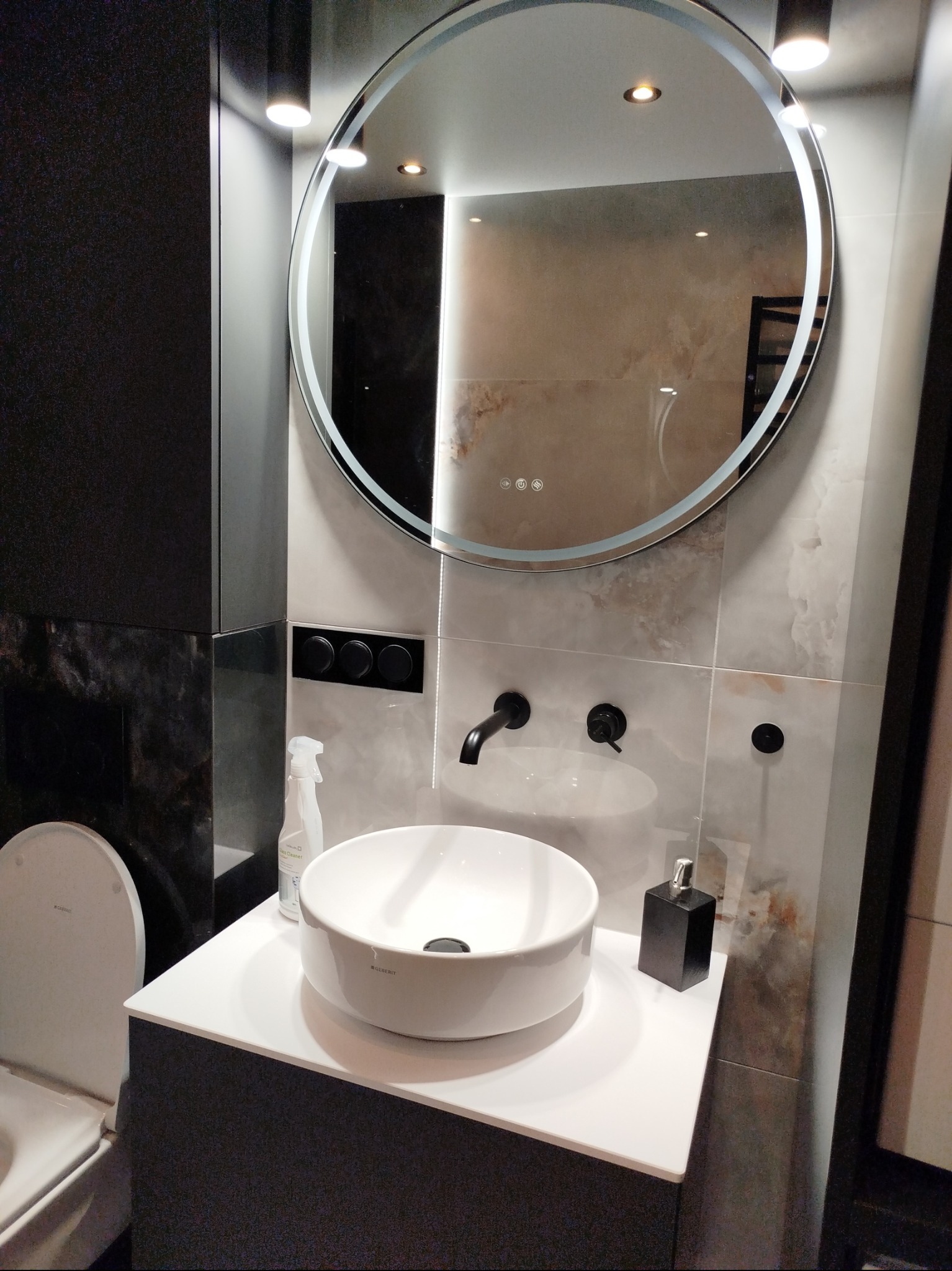 Nowoczesna łazienka z okrągłym lustrem LED, białą umywalką nablatową i czarną armaturą. Marmurowe płytki na ścianach. Widoczny fragment toalety i akcesoria.