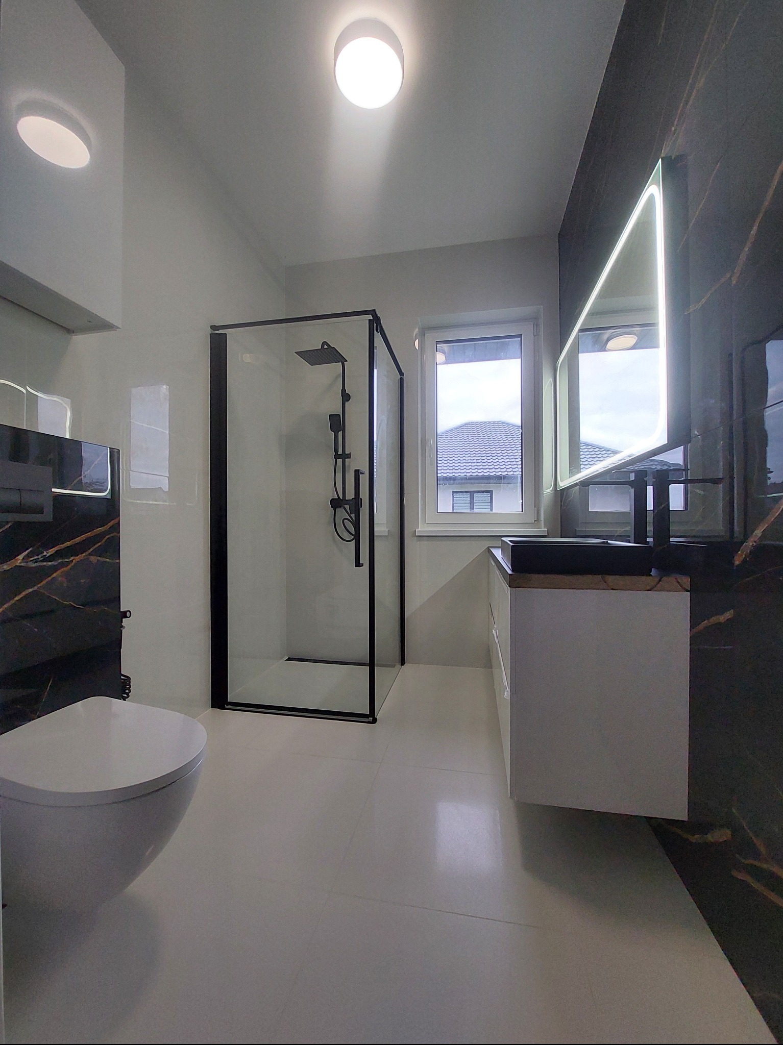 Nowoczesna łazienka po remoncie: kabina prysznicowa z czarną ramą, podświetlane lustro nad umywalką i marmurowe płytki. Minimalistyczny design, jasne płytki podłogowe.