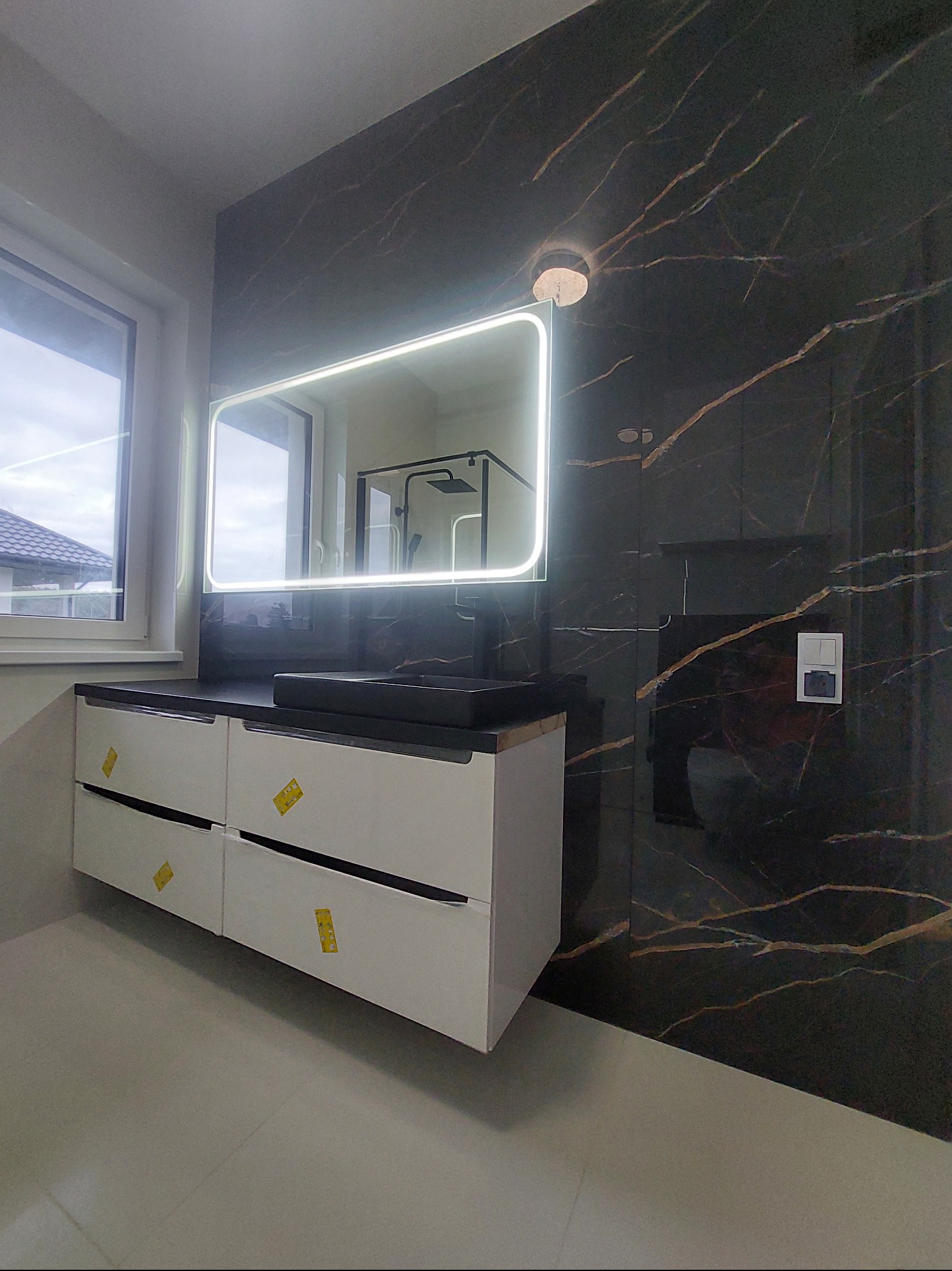 Nowoczesna łazienka z czarną umywalką i podświetlanym lustrem. Ściana wyłożona płytkami imitującymi marmur z złotymi żyłami. Białe szafki pod umywalką z czarnymi uchwytami.