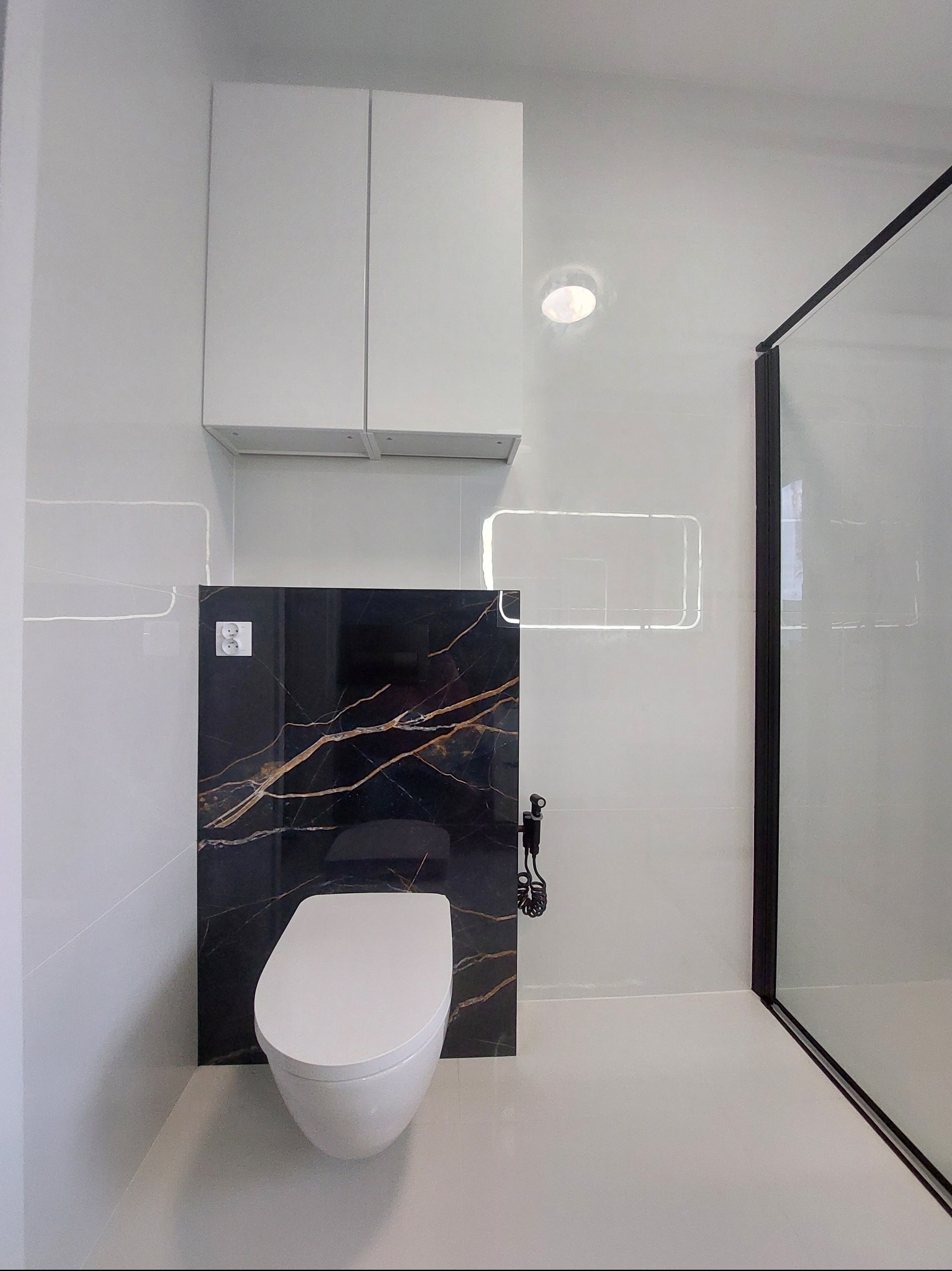 Nowoczesna łazienka z podwieszaną toaletą na tle czarnej płyty z żyłkowaniem, białe płytki, szafka wisząca i kabina prysznicowa z czarnymi profilami.