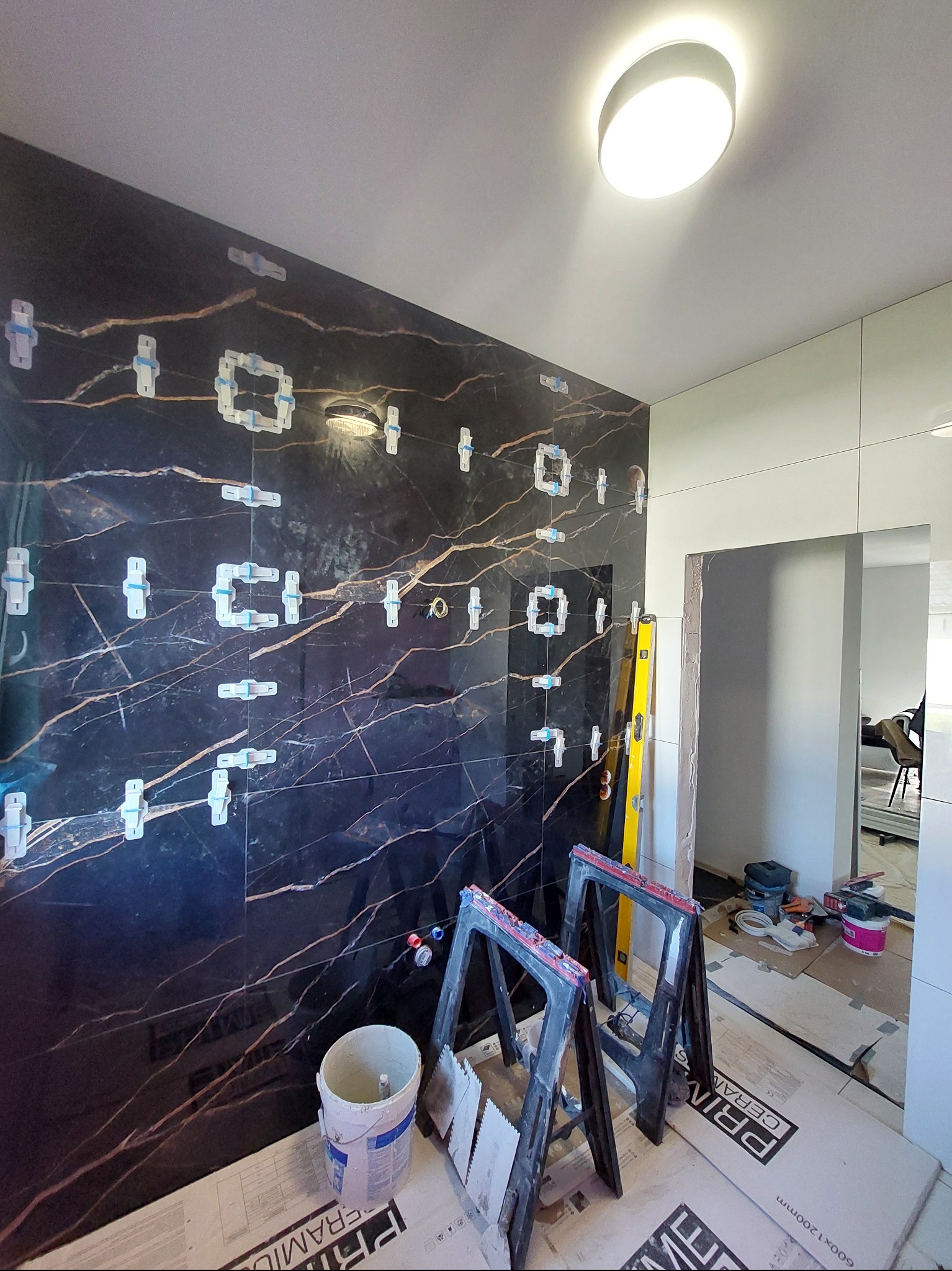 Częściowo wyłożona ściana czarnymi płytkami imitującymi marmur z widocznymi krzyżykami dystansowymi, drabina i wiadro z zaprawą w trakcie prac wykończeniowych.
