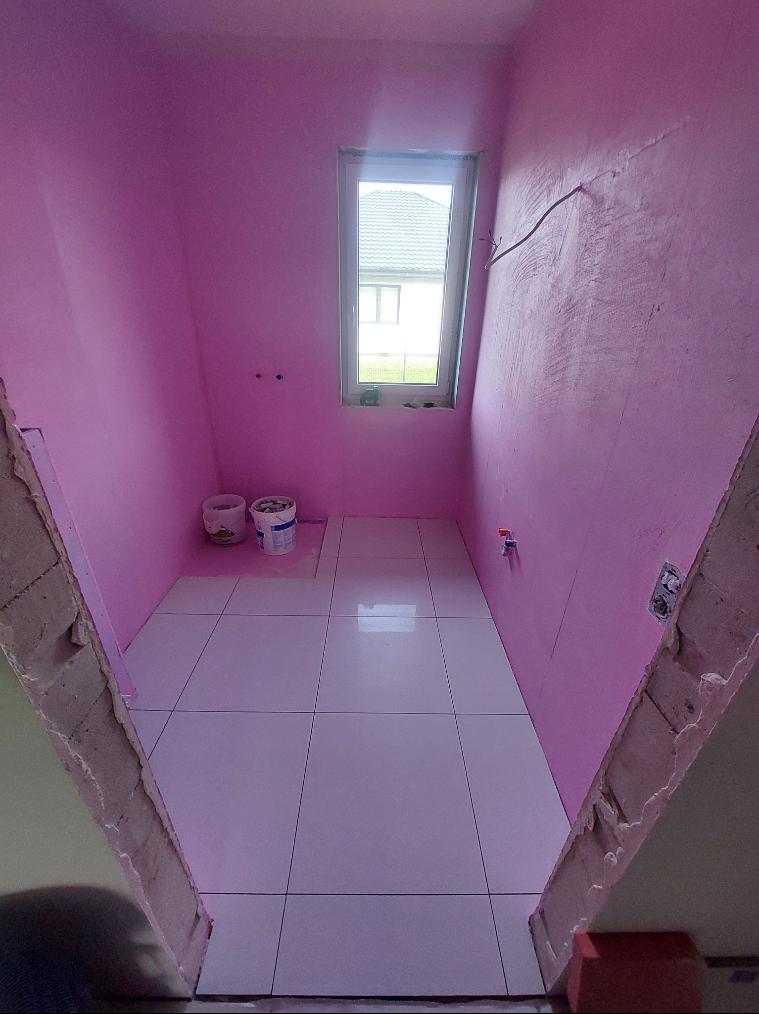 Różowa łazienka w trakcie prac: białe płytki podłogowe, ściany pomalowane na różowo, widoczne puszki z farbą i fragment okna z widokiem na dom.