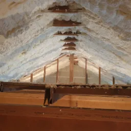 Poddasze z drewnianą konstrukcją krokwiową pokryte grubą warstwą białej, spienionej izolacji natryskowej. Widoczne nierówności i struktura piany.