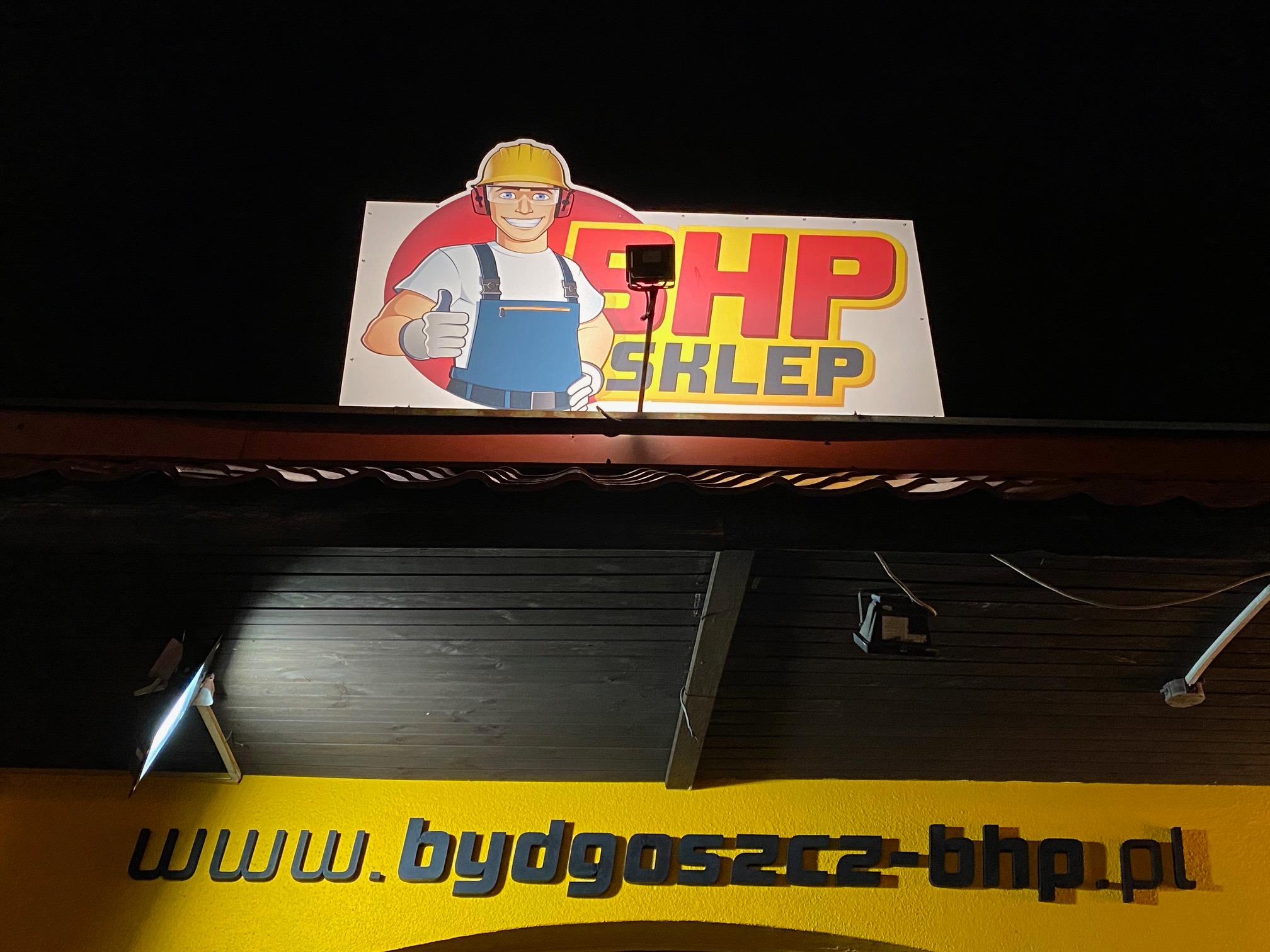 Podświetlany szyld sklepu BHP nocą z rysunkową postacią w kasku i kombinezonie oraz adresem strony internetowej.