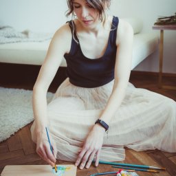 Sylwia Kos Fotografia - Kobieta w czarnym topie i tiulowej spódnicy siedzi na podłodze, miesza farby na palecie za pomocą pędzla. Obok leżą tubki z farbami i pojemnik z wodą.