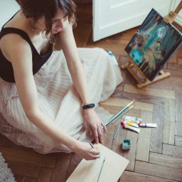 Sylwia Kos Fotografia - Kobieta siedzi na podłodze z parkietu w sukience z tiulu, malując na drewnianej palecie. Na sztaludze widnieje abstrakcyjny obraz. Obok leżą pędzle i farby.