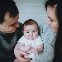 Sylwia Kos Fotografia - Rodzice spoglądają z czułością na niemowlę ubrane w białe body z delikatnym kwiatowym haftem, trzymane na rękach, na tle białej ceglanej ściany.