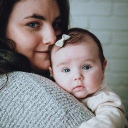 Sylwia Kos Fotografia - Portret matki trzymającej na ramieniu niemowlę z kokardką we włosach, na tle ściany z cegieł.