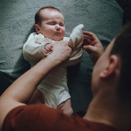 Sylwia Kos Fotografia - Noworodek w białym body, leżący na szarym kocu, ubierany przez osobę w czerwonobrązowej koszulce, ujęcie z góry.