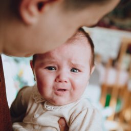 Sylwia Kos Fotografia - Niemowlę z grymasem na twarzy, trzymane przez osobę dorosłą, ubrane w kremową sukienkę z koronkowym kołnierzykiem.