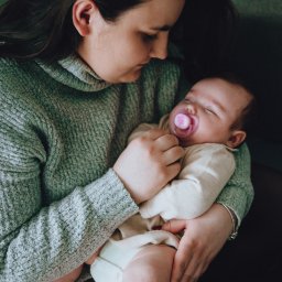 Sylwia Kos Fotografia - Mama w szarym swetrze trzyma śpiące niemowlę z różowym smoczkiem w jasnym ubranku.