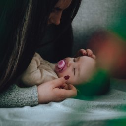 Sylwia Kos Fotografia - Matka czule spogląda na śpiące niemowlę z różowym smoczkiem, leżące na miękkim, błękitnym kocyku, z dłońmi matki delikatnie ułożonymi na jego główce, w tle szara ściana.