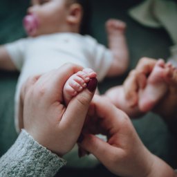 Sylwia Kos Fotografia - Mama trzyma stópkę noworodka z bordowym lakierem na paznokciach, w tle śpiące dziecko z smoczkiem.