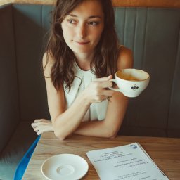 Sylwia Kos Fotografia - Kobieta z brązowymi włosami siedzi w kawiarni, trzymając filiżankę z napojem, obok leży spodek i kartki z tekstem.