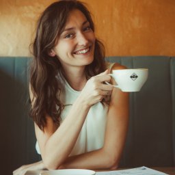 Sylwia Kos Fotografia - Uśmiechnięta kobieta z filiżanką kawy w kawiarni, siedząca przy drewnianym stole z menu i talerzykiem, oświetlona ciepłym światłem.