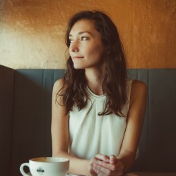 Sylwia Kos Fotografia - Kobieta siedzi przy stoliku w kawiarni, zamyślona, obok filiżanka kawy z logo na tle złotej ściany i szarej sofy.