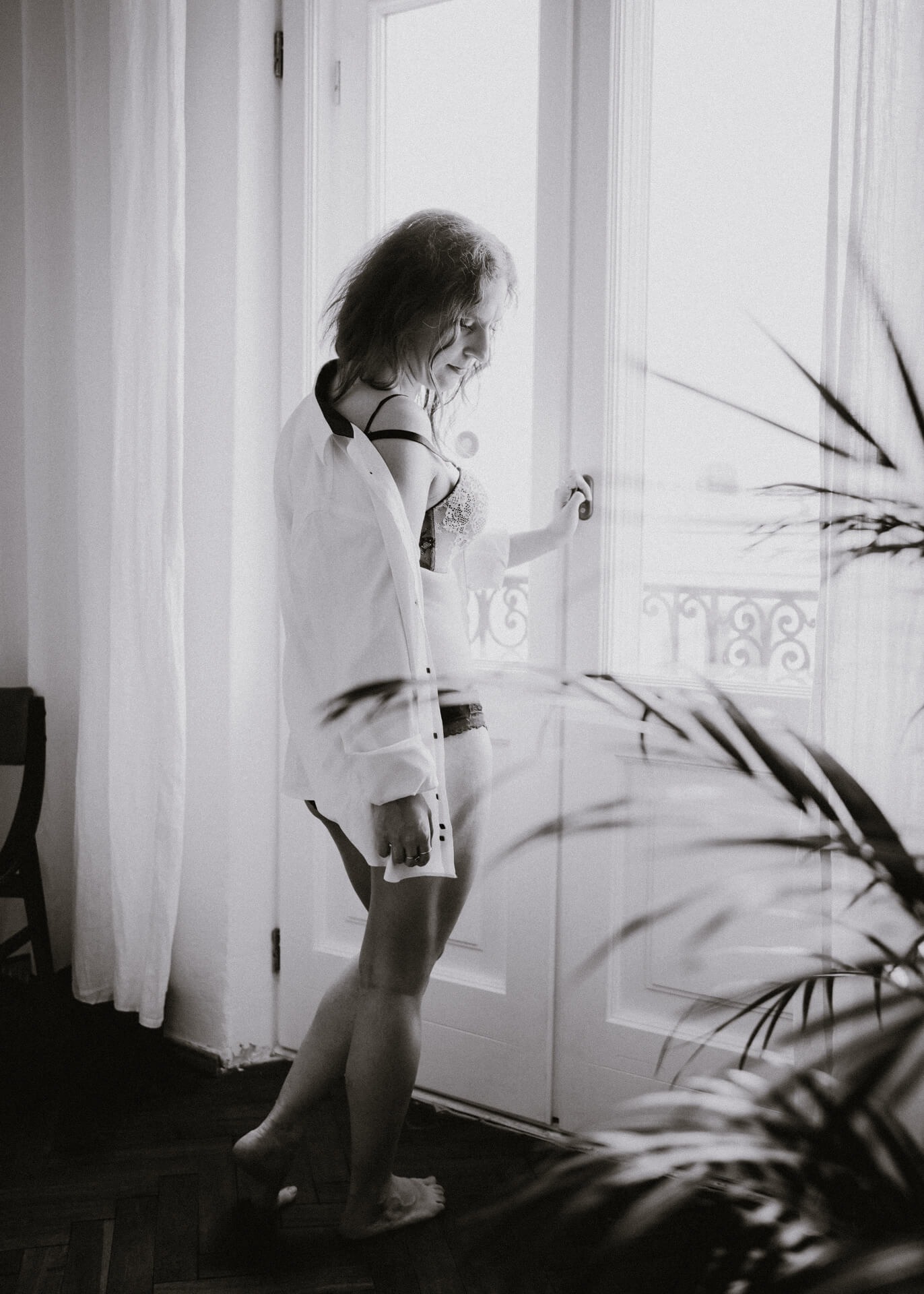 Czarno-białe zdjęcie kobiety w bieliźnie i rozpiętej koszuli, stojącej przy przeszklonych drzwiach balkonowych z widokiem na balkon z ozdobną balustradą, częściowo zasłoniętej rośliną doniczkową.