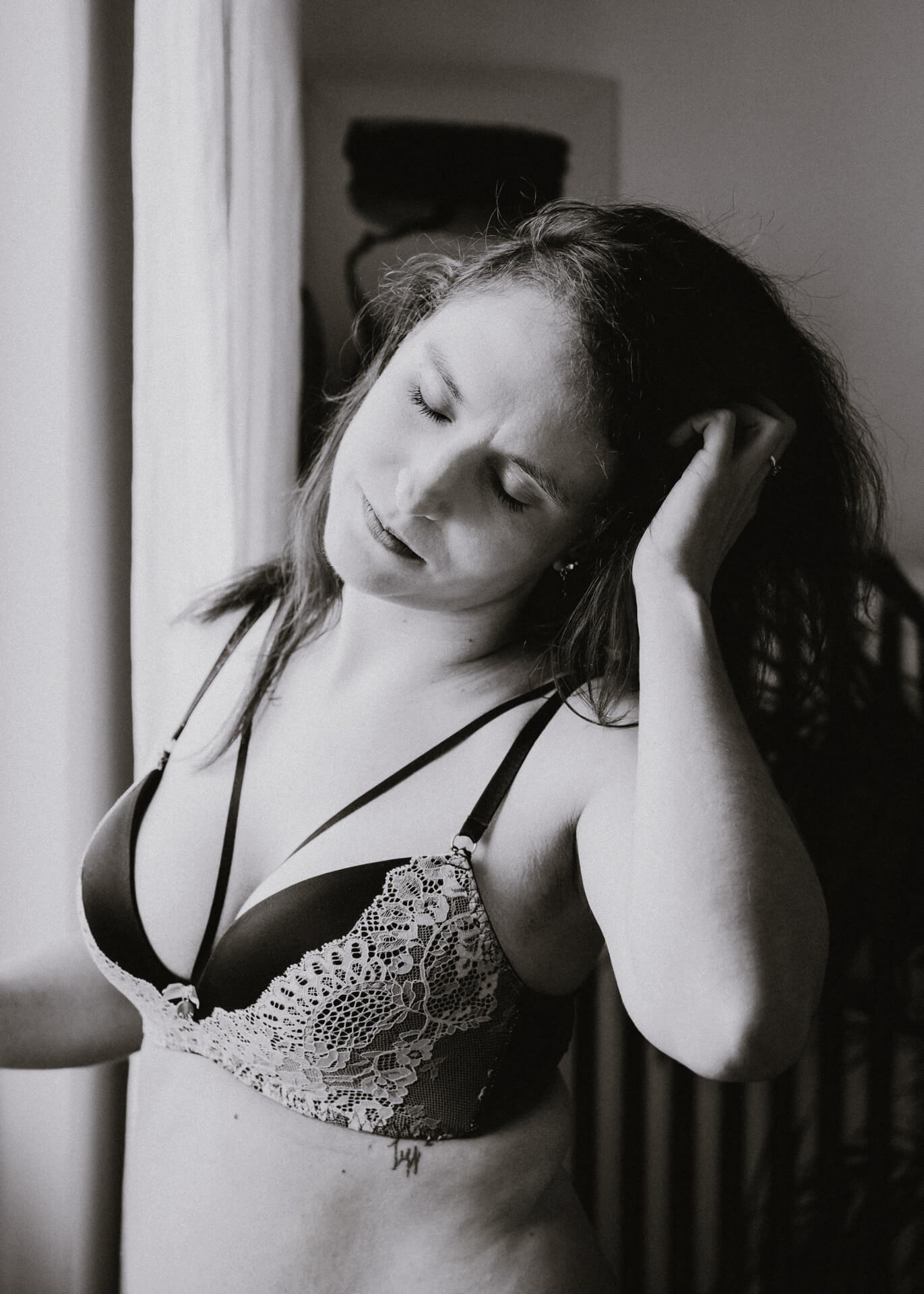 Czarno-białe zbliżenie kobiety w koronkowej bieliźnie, stojącej przy oknie i dotykającej włosów, z widocznym tatuażem na brzuchu.
