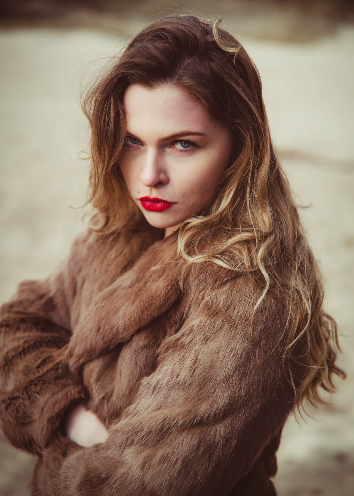 Portret kobiety w brązowym futrze z intensywnie czerwoną szminką, patrzącej z góry z założonymi rękami na neutralnym tle.