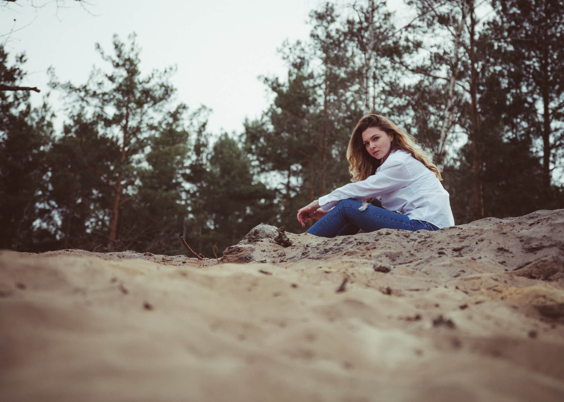 Kobieta w białej koszuli i dżinsach siedzi na piaszczystym wzgórzu z lasem sosnowym w tle, patrząc w obiektyw.