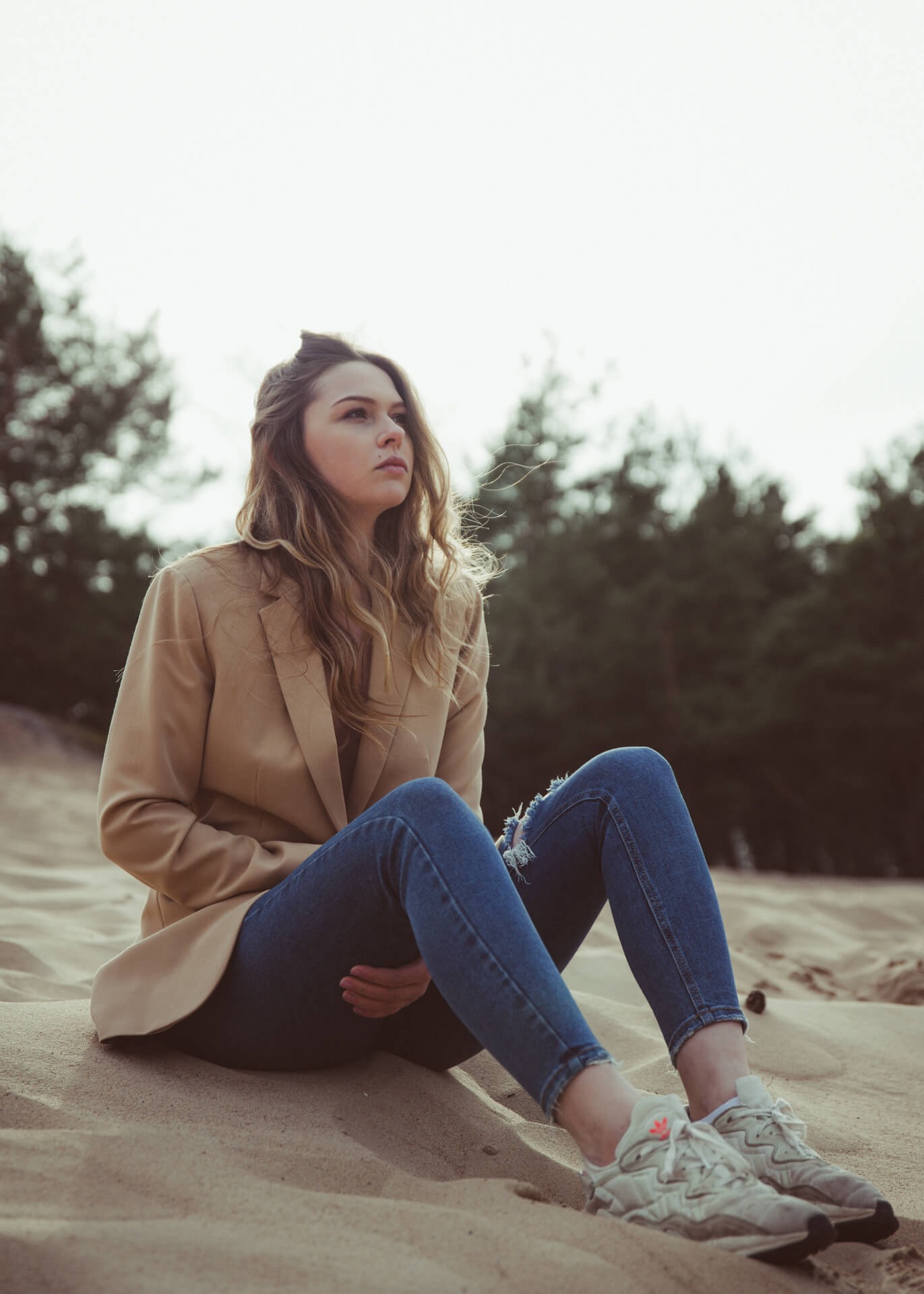 Młoda kobieta w beżowej marynarce i podartych jeansach siedzi na piaszczystej wydmie, zamyślona spoglądając w dal, z lasem w tle.