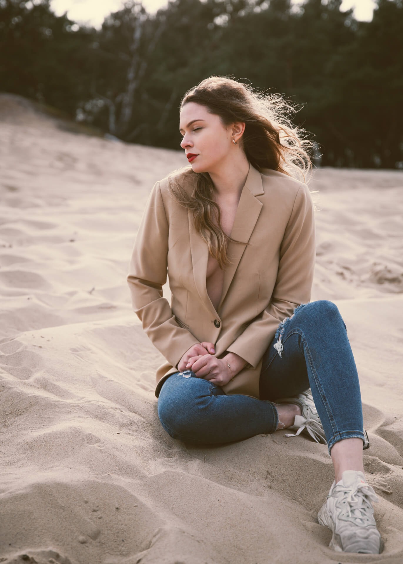 Portret kobiety w beżowej marynarce i podartych jeansach siedzącej na piaszczystej wydmie z lasem w tle, włosy rozwiane na wietrze, zamyślony wyraz twarzy.