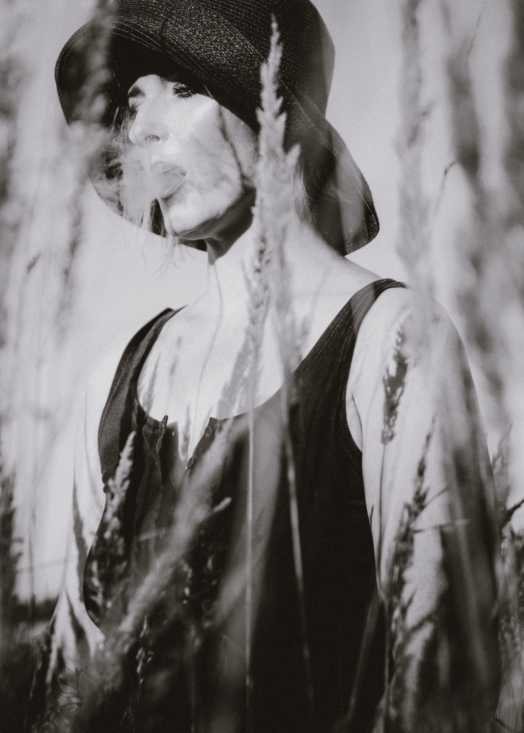 Portret kobiety w kapeluszu, zarys sylwetki w plenerze, czarno-biała fotografia z elementami trawy na pierwszym planie, artystyczne ujęcie.