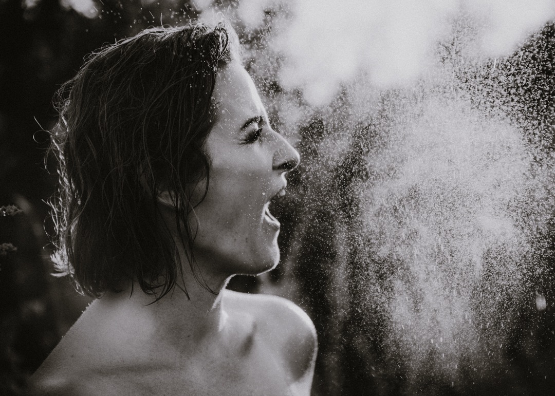 Czarno-białe zdjęcie kobiety z otwartymi ustami, spryskiwanej wodą, profil, oświetlenie od tyłu.