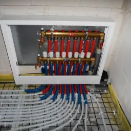 Rozdzielacz ogrzewania podłogowego w białej szafce, z widocznymi rurami w kolorach czerwonym i niebieskim, ułożonymi na siatce podłogowej.