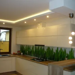 Nowoczesna kuchnia z białymi, lakierowanymi szafkami, drewnianym blatem i panelem szklanym z motywem trawy, oświetlona lampami sufitowymi i wiszącą lampą z trzema kulami.