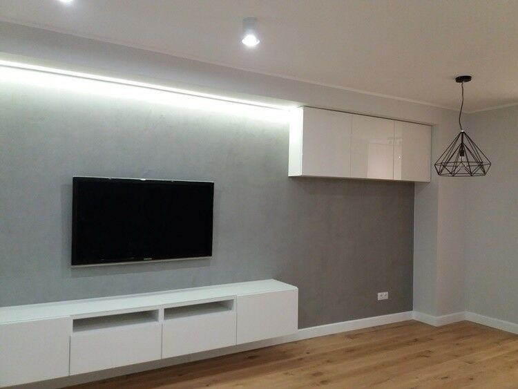 Nowoczesny salon z szarą ścianą, białymi meblami RTV i wiszącymi szafkami, drewnianą podłogą oraz geometryczną lampą wiszącą.