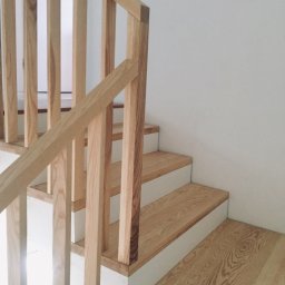 Interwald - Drewniane schody o jasnym odcieniu drewna, z białymi podstopnicami, widziane z dołu, z minimalistyczną balustradą z pionowych szczebli.