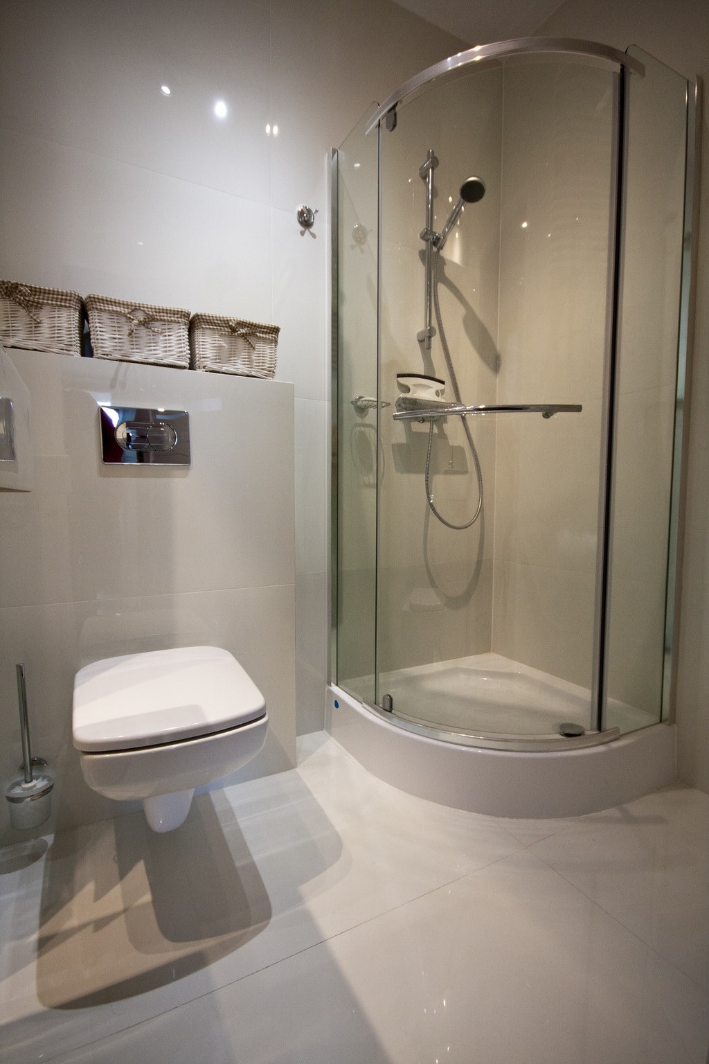 Jasna łazienka z kabiną prysznicową narożną, podwieszaną toaletą z chromowanym przyciskiem spłukującym i trzema wiklinowymi koszykami na ścianie.