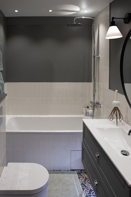 Nowoczesna łazienka z grafitową ścianą, jasnymi płytkami ułożonymi pionowo, białą wanną i umywalką na szafce w kolorze grafitu, okrągłym lustrem oraz dekoracyjnym oświetleniem i armaturą.