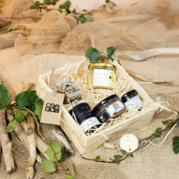 Ręcznie robiony, drewniany kosz prezentowy wypełniony słoiczkami z miodem, konfiturami i smalcem, ozdobiony jutowym materiałem, gałązkami bluszczu i drewnianymi elementami dekoracyjnymi.