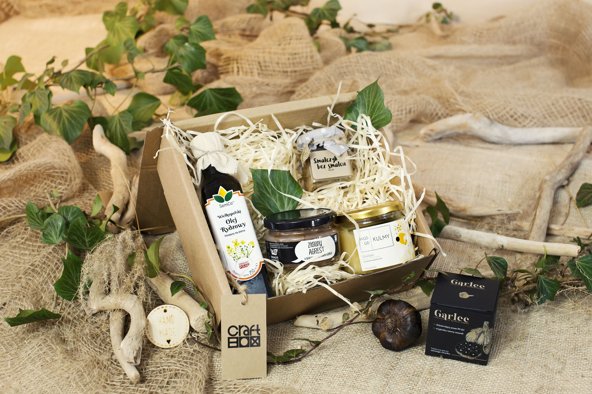 Ozdobny kartonowy kosz prezentowy wypełniony słoiczkami z miodem, smalcem wegańskim, olejem rydzowym i czarnym czosnkiem, udekorowany jutą, drewnem i bluszczem.