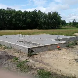 Wylany fundament pod budynek z widocznymi elementami instalacji, na zewnątrz teren budowy i zieleń.