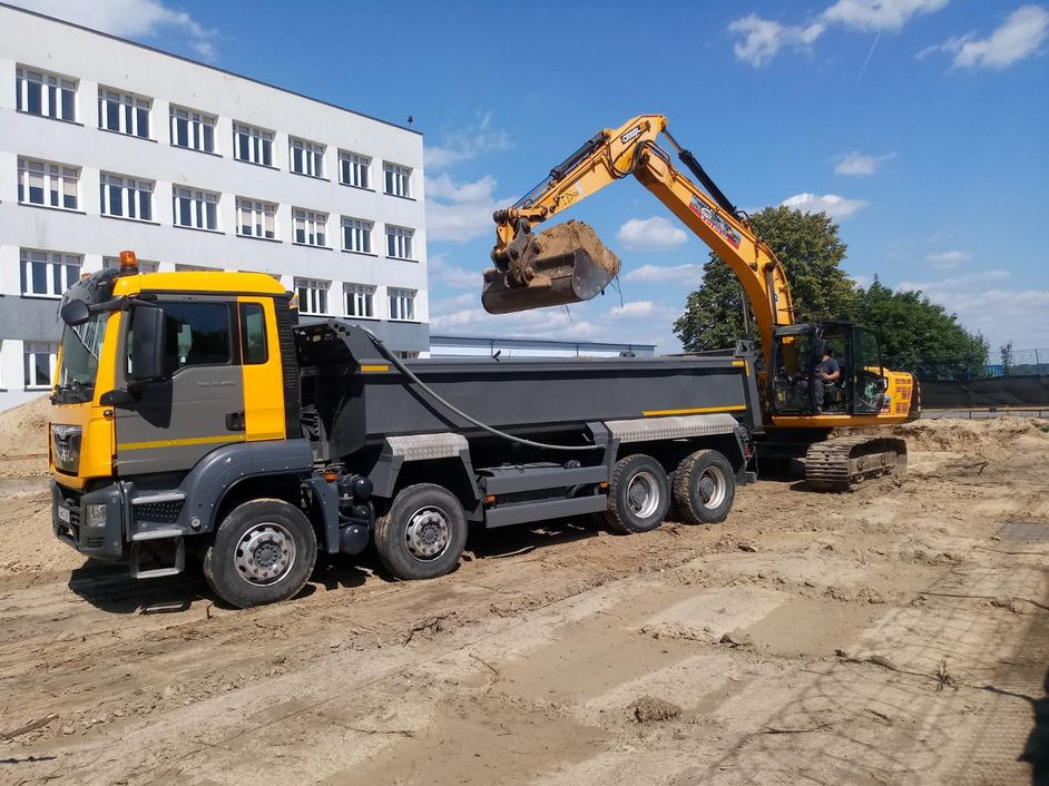 Żółto-szara ciężarówka MAN i żółta koparka Hyundai na placu budowy podczas załadunku ziemi, w tle budynek z białej cegły i błękitne niebo.