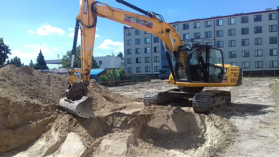 Żółta koparka gąsienicowa z łyżką zagłębia się w piaszczysty wykop, budynek mieszkalny w tle.