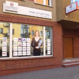 Fasada budynku z cegły z szyldem 'Dom Ubezpieczeniowy Spectrum' i napisem 'twoje ubezpieczenia'. W oknie naklejki z logotypami różnych towarzystw ubezpieczeniowych.