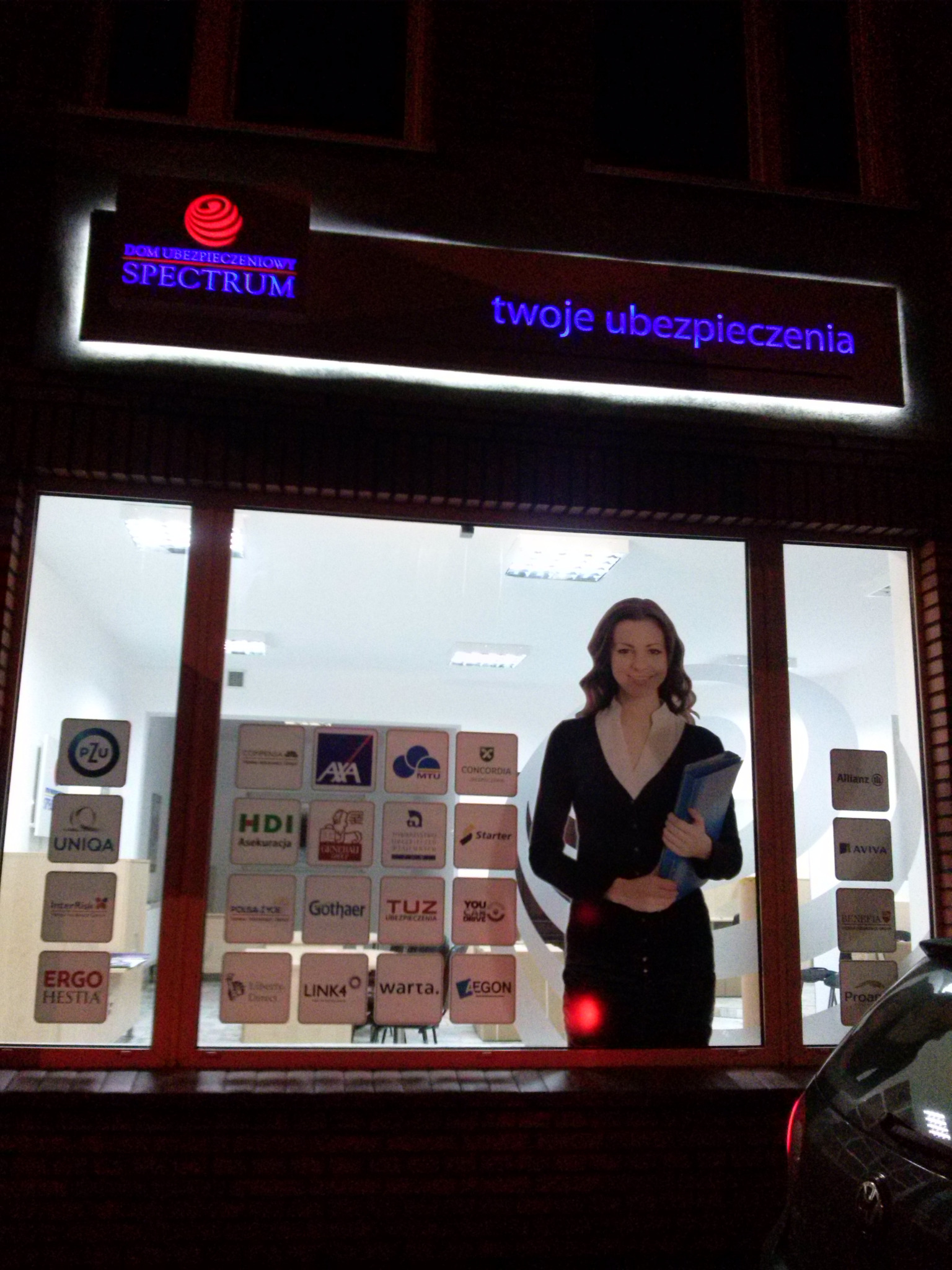 Fasada budynku agencji ubezpieczeniowej z podświetlanym szyldem 'DOM UBEZPIECZENIOWY SPECTRUM' i napisem 'twoje ubezpieczenia', w oknie naklejki z logotypami różnych towarzystw ubezpieczeniowych...