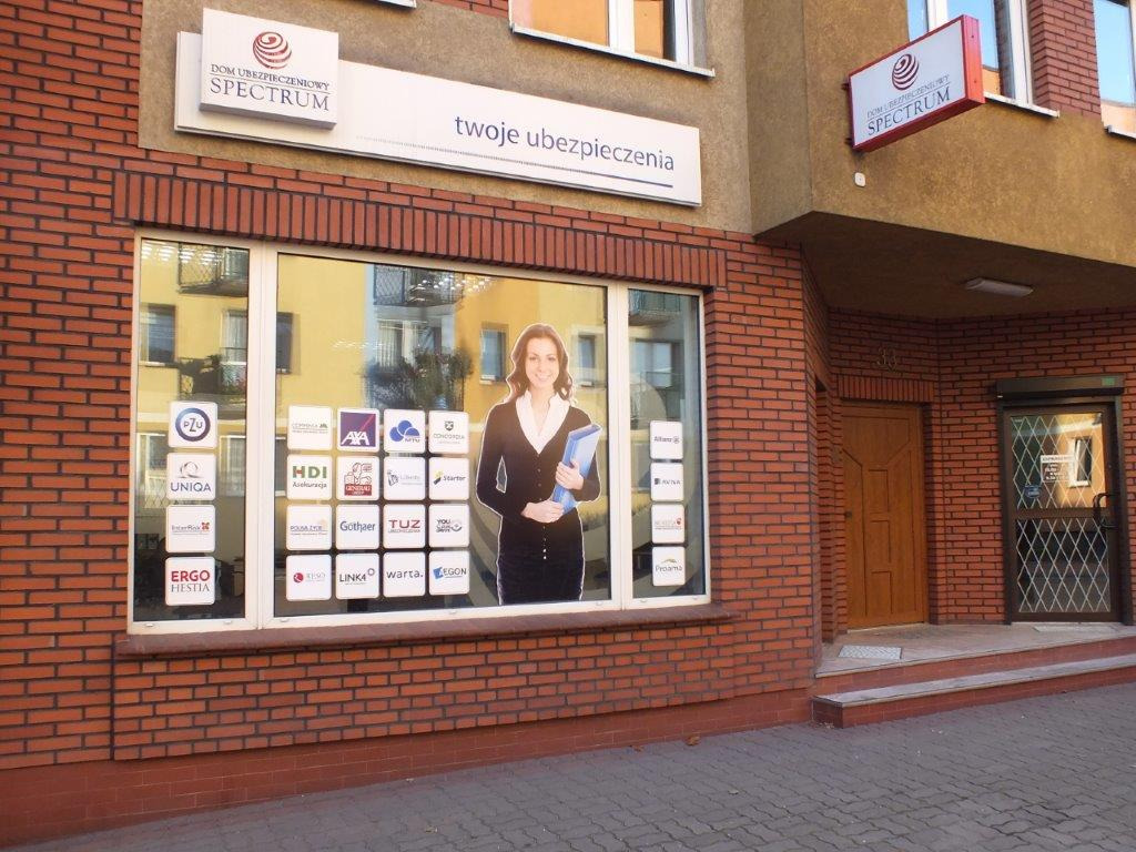 Fasada budynku z cegły z szyldem 'Dom Ubezpieczeniowy Spectrum' i napisem 'twoje ubezpieczenia'. W oknie naklejki z logotypami różnych towarzystw ubezpieczeniowych.