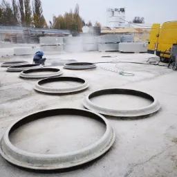 Pracownik w niebieskim kombinezonie czyści powierzchnię betonowych pierścieni na zewnątrz, obok żółtego busa z urządzeniem do obróbki strumieniowo-ściernej.